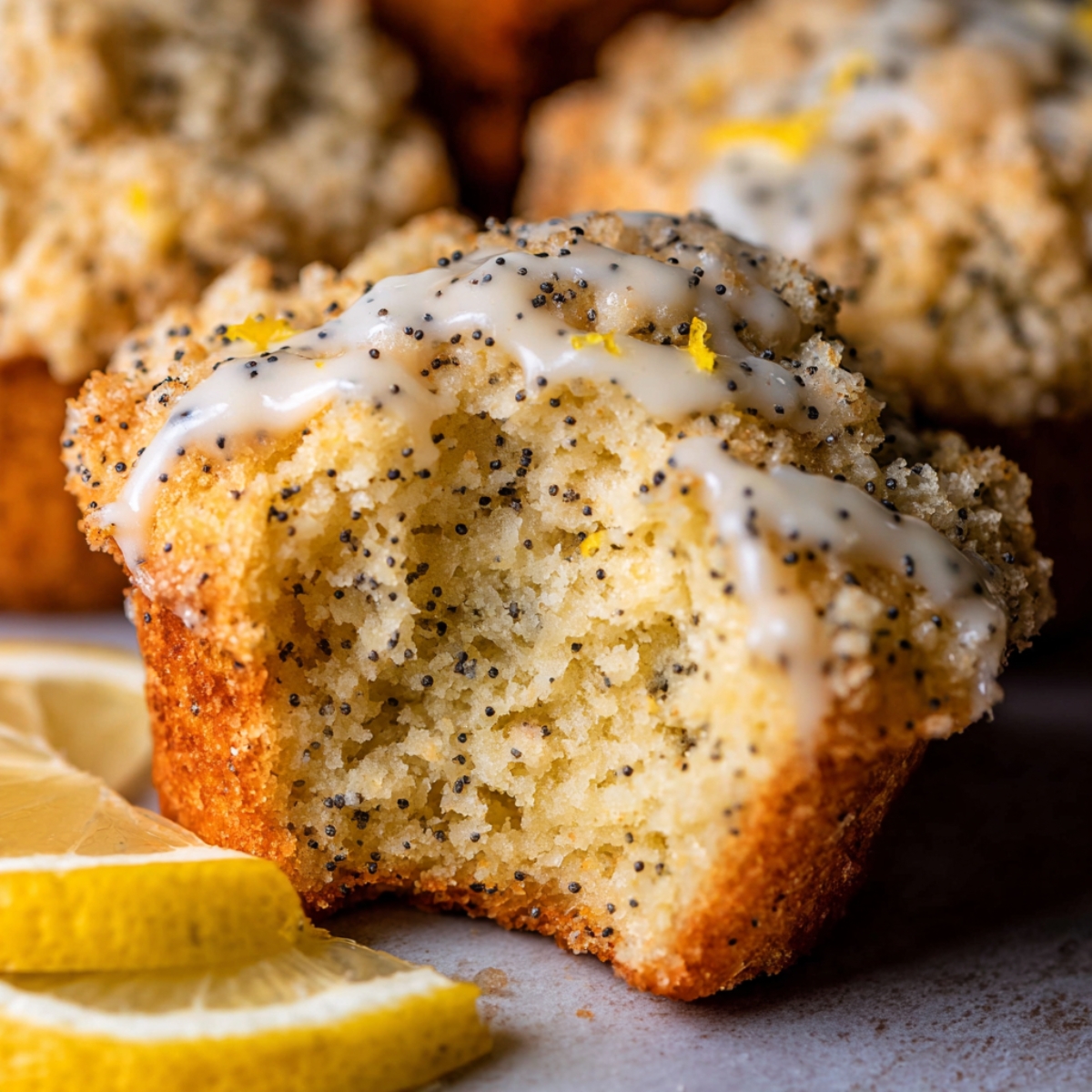 A close-up of a Lemon Poppy Seed Muffins with a bite taken out, showcasing a fluffy interior, poppy seeds, and a crumbly topping with icing drizzle, accompanied by lemon slices.