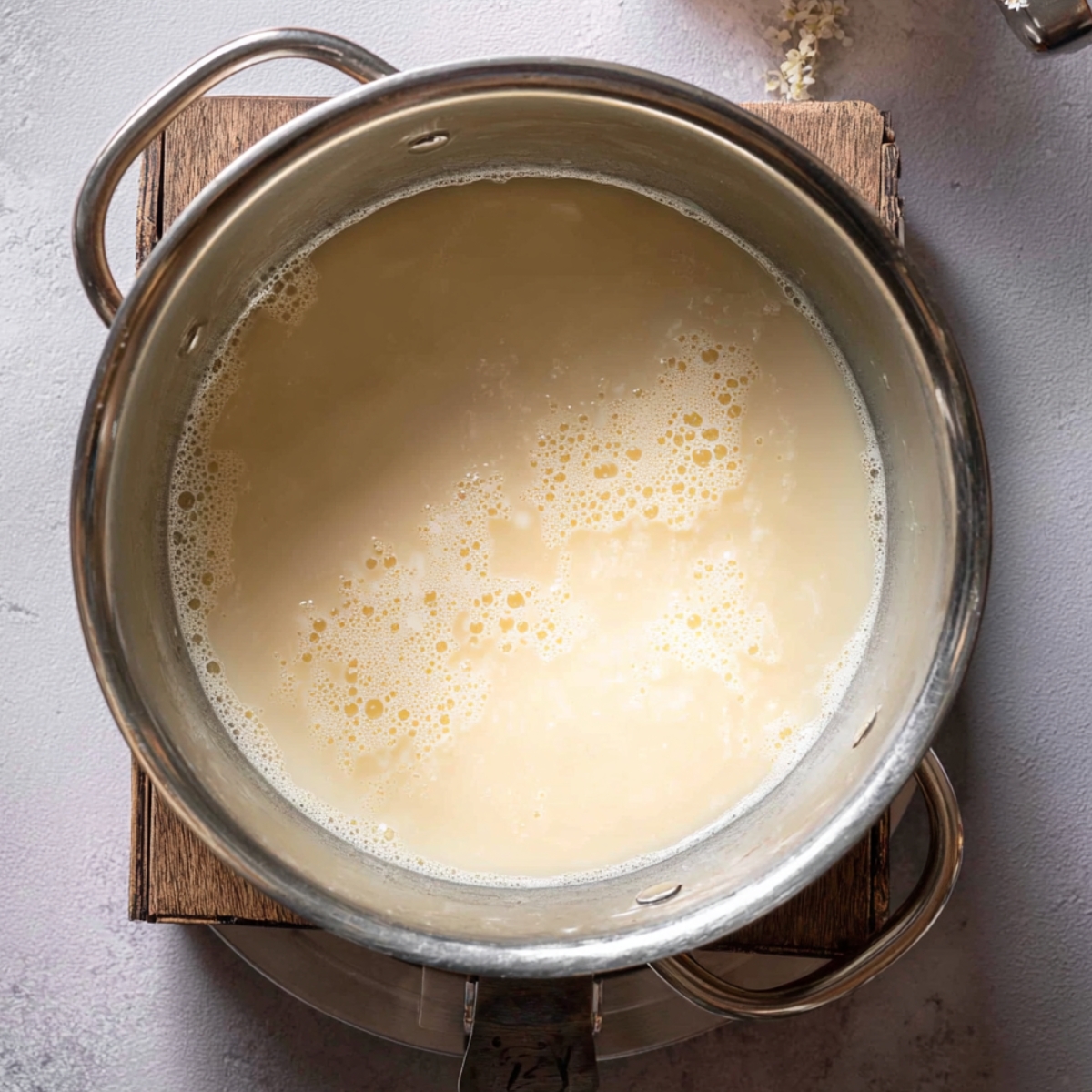 Homemade White Chocolate Cupcakes 11 Close-up of a metal bowl with a pale white chocolate mixture, slightly bubbly, on a wooden surface with white chocolate pieces scattered nearby.