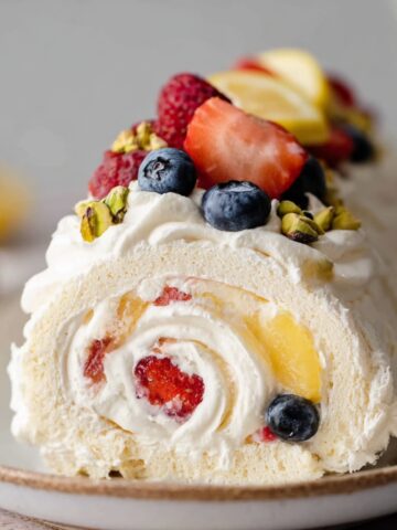 A beautifully rolled gluten-free Meringue Roulade topped with fresh strawberries, blueberries, pistachios, and lemon slices, showcasing a creamy vanilla filling inside.