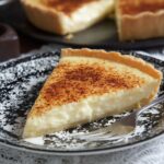A slice of freshly baked Milk Tart with a creamy custard filling, topped with a sprinkle of cinnamon. The tart is served on a decorative black-and-white plate, with the whole tart in the background.