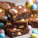 A stack of Mini Egg Brownies with visible chunks of white chocolate and colorful mini eggs, placed on a table with more mini eggs scattered around.