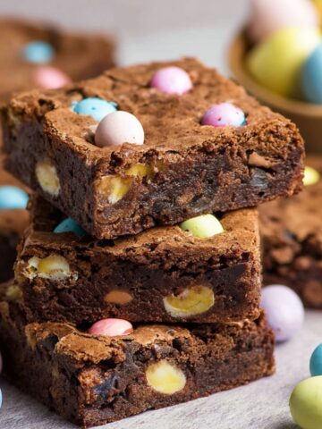 Easy Mini Egg Brownies Recipe 5 A stack of Mini Egg Brownies with visible chunks of white chocolate and colorful mini eggs, placed on a table with more mini eggs scattered around.