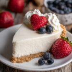 A slice of No Bake Cheesecake with a heart-shaped strawberry, blueberries, and whipped cream on top, served on a white plate.