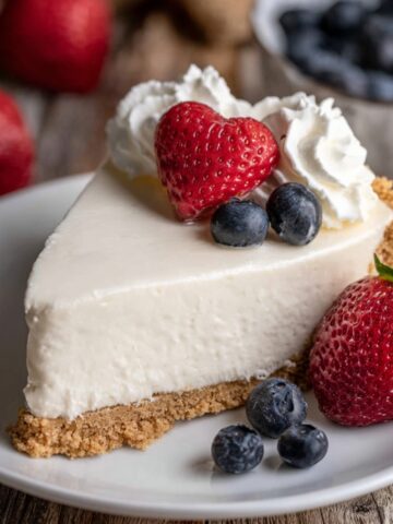 A slice of No Bake Cheesecake with a heart-shaped strawberry, blueberries, and whipped cream on top, served on a white plate.