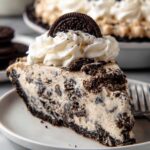 A slice of Oreo pie with creamy filling, topped with whipped cream and a whole Oreo cookie, served on a white plate.