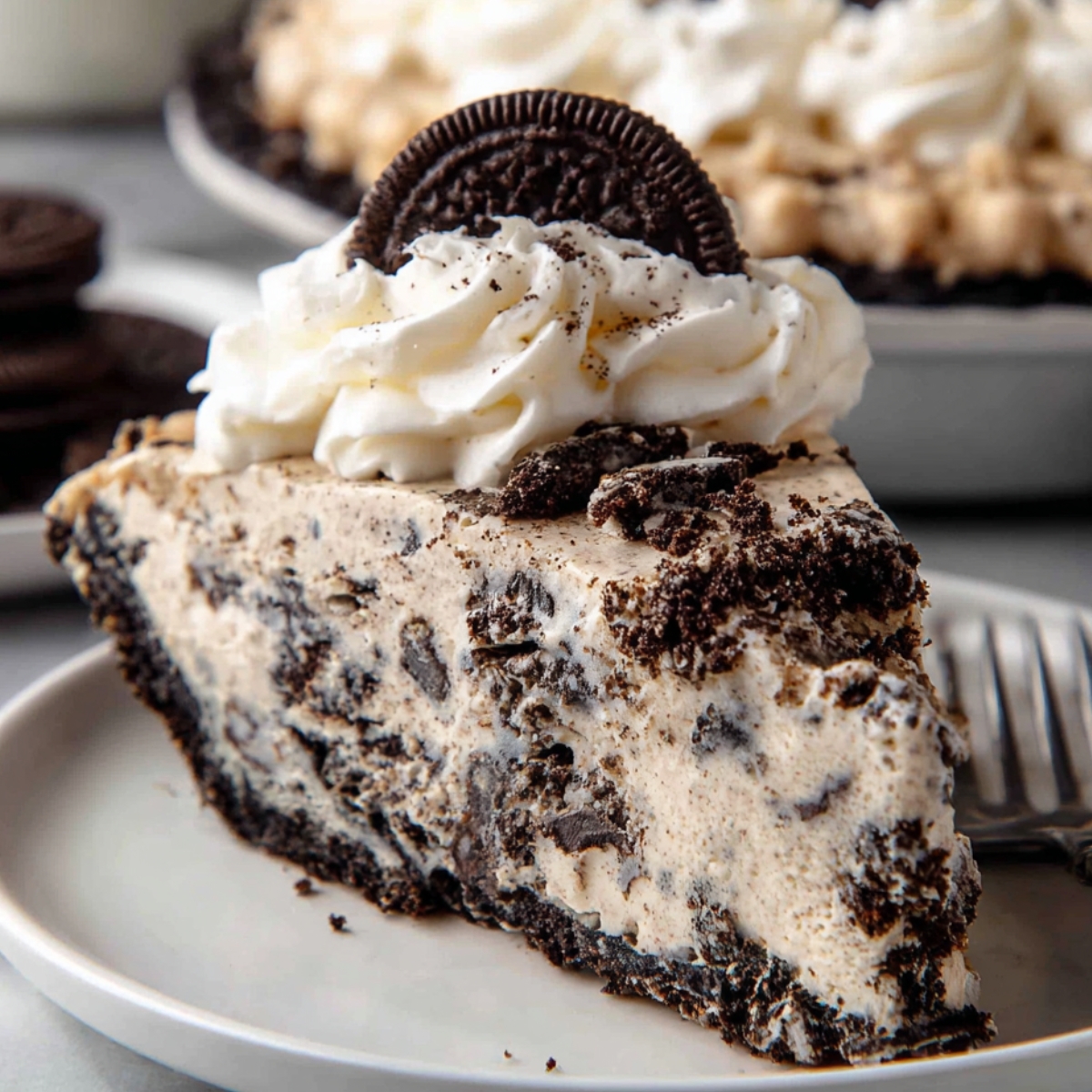 A slice of Oreo pie with creamy filling, topped with whipped cream and a whole Oreo cookie, served on a white plate.