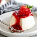A beautifully plated Panna Cotta topped with vibrant strawberry sauce and a fresh strawberry garnish, with a spoon resting beside it.