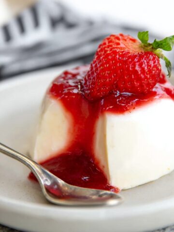 Classic Panna Cotta Recipe 1 A beautifully plated Panna Cotta topped with vibrant strawberry sauce and a fresh strawberry garnish, with a spoon resting beside it.