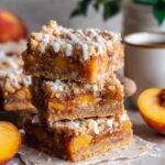 A stack of freshly baked Peach Cobbler Bars with a golden crumble topping and sweet peach filling, drizzled with icing for extra sweetness.