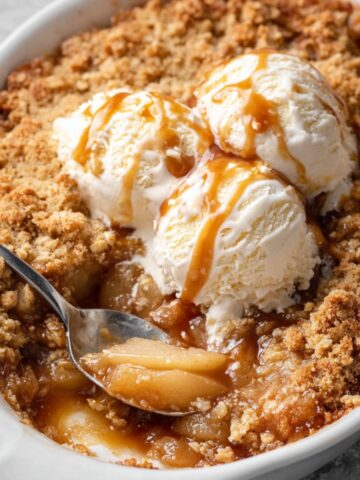 The Best Pear Crumble Recipe 2 A bowl filled with baked Pear Crumble, topped with two scoops of vanilla ice cream, drizzled with caramel sauce, and a spoonful of the warm fruit filling is scooped out.
