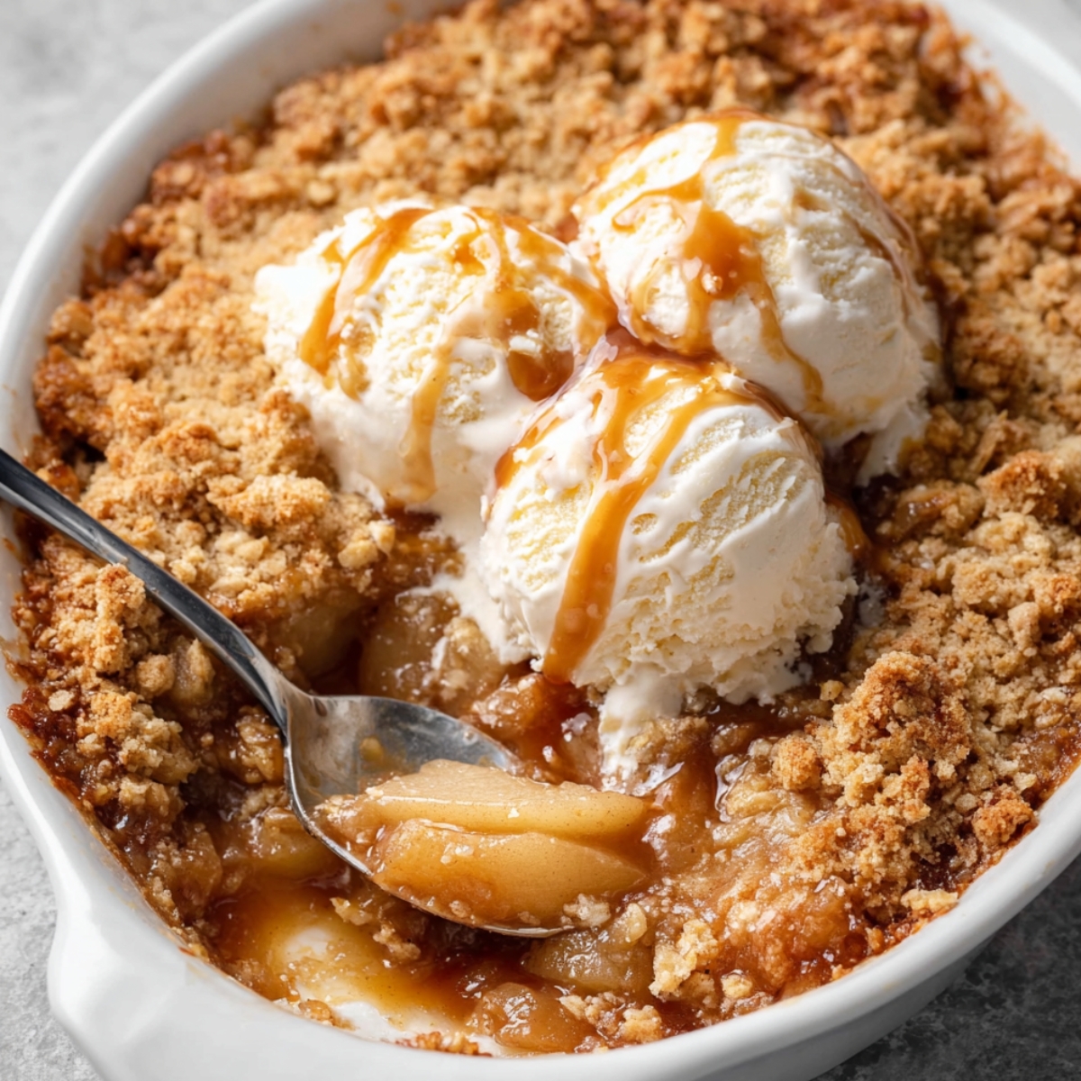 A bowl filled with baked Pear Crumble, topped with two scoops of vanilla ice cream, drizzled with caramel sauce, and a spoonful of the warm fruit filling is scooped out.
