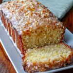 A close-up of a freshly baked loaf of Pineapple Coconut Bread with a glossy glaze and toasted coconut flakes on top. A slice has been cut, showing the soft interior.