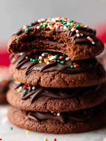Easy Piped Chocolate Butter Cookies 1 A stack of Piped Chocolate Butter Cookies drizzled with dark chocolate and sprinkled with festive red, green, and white sprinkles. One cookie has a bite taken out to reveal its soft texture.