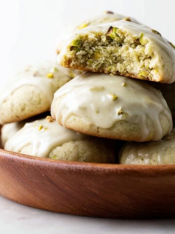 Best Pistachio Drop Cookies Recipe 3 A close-up of soft Pistachio Drop Cookies topped with a smooth, glossy glaze. The cookies are arranged in a wooden bowl, with one cookie broken in half to reveal the soft, pistachio-filled interior.