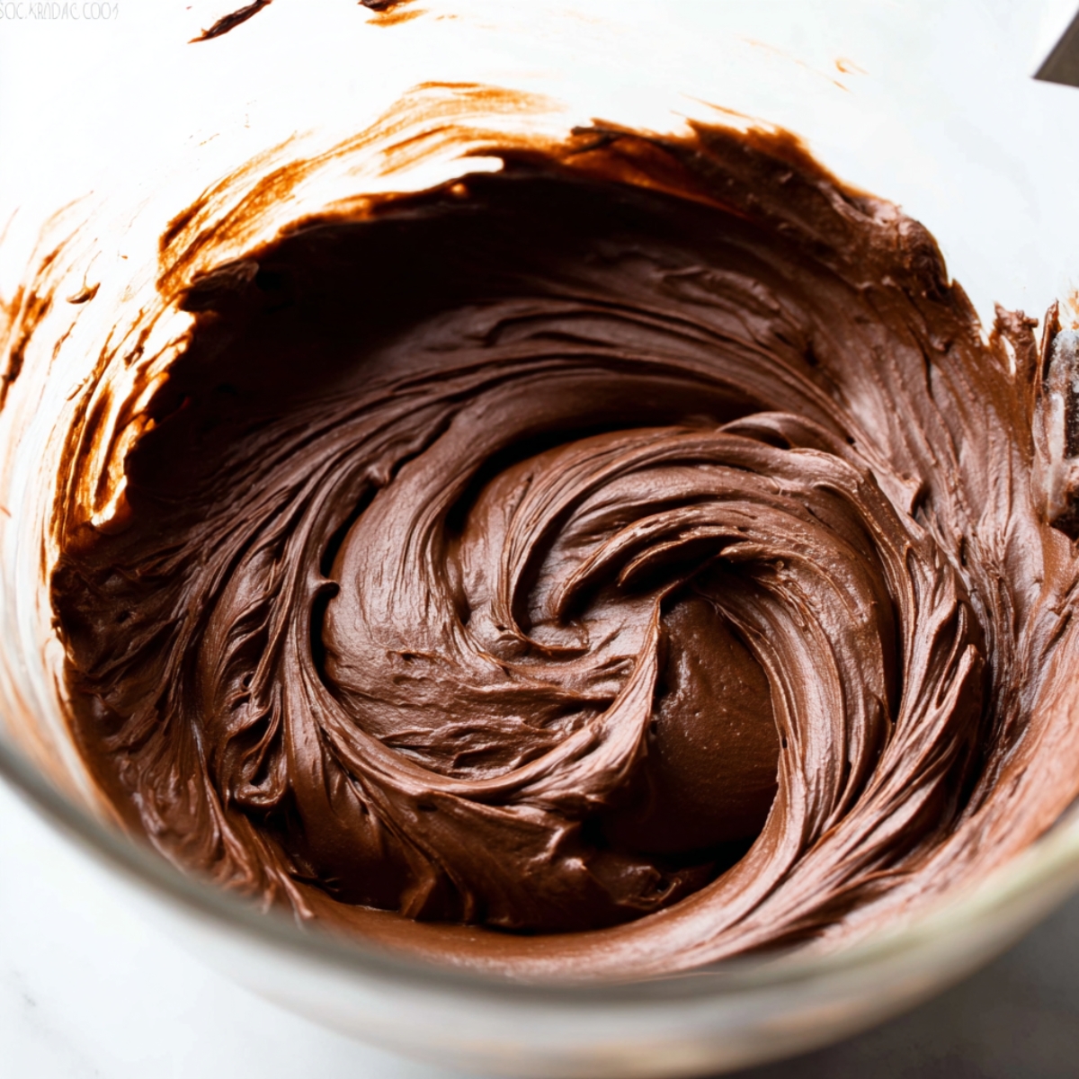 Deliciously Moist Chocolate Layer Cake 11 A close-up of smooth chocolate frosting being mixed in a glass bowl, with soft, glossy swirls creating a rich, velvety texture.