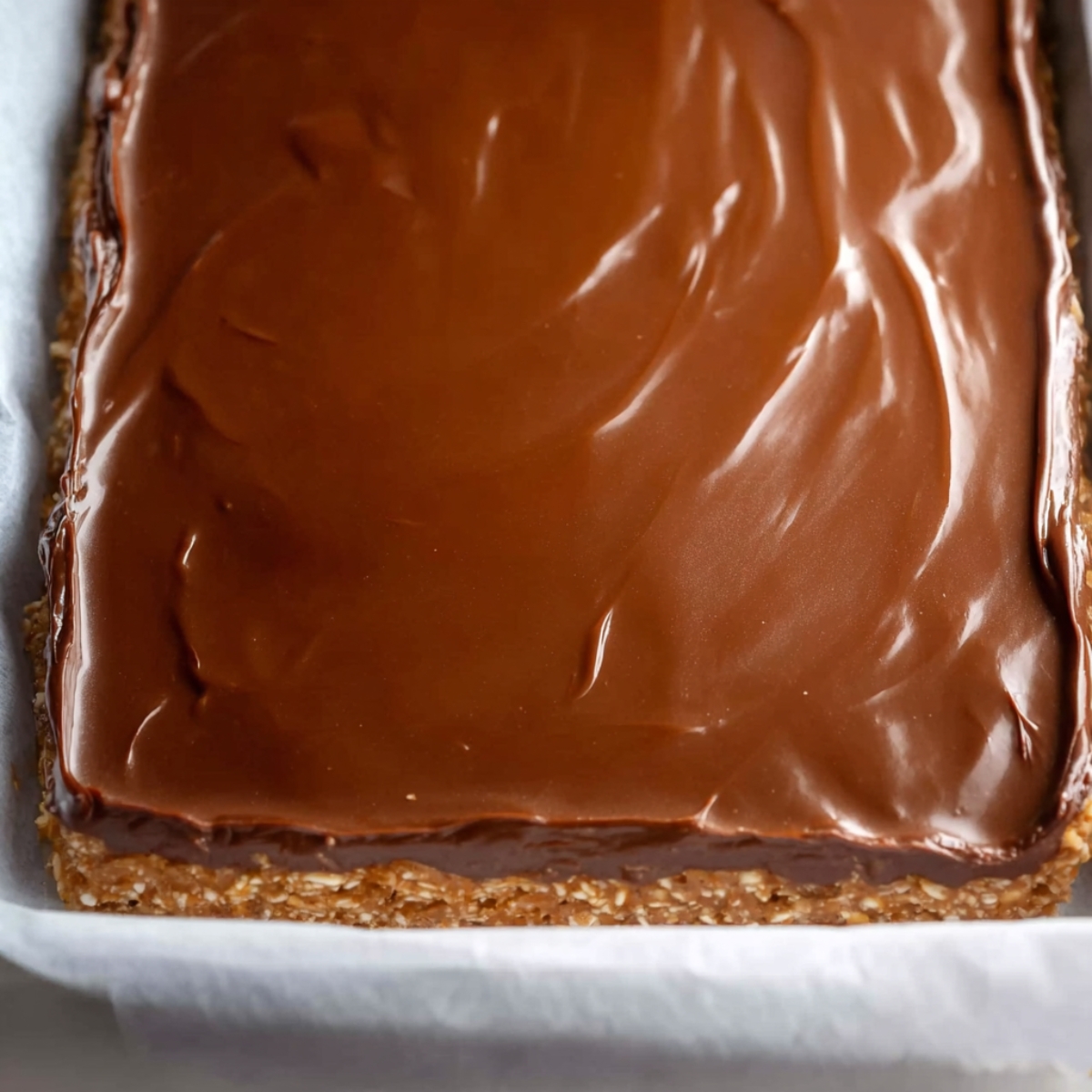 Healthy Nutty Chocolate Bars Recipe 11 A square pan of oat bars with a smooth, glossy chocolate layer spread evenly on top, ready to set.