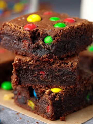 Easy Protein Brownies with M&Ms Recipe 1 stack of fudgy Protein Brownies with M&Ms visible on top and inside. The brownies have a rich, dark chocolate texture, and the bright M&M's add a playful pop of color to the dessert.