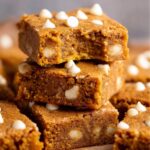 A stack of warm, freshly baked Pumpkin Blondies with a bite taken out of the top one, revealing its soft, chewy interior and white chocolate chips.