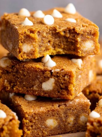 The Best Pumpkin Blondies Recipe 1 A stack of warm, freshly baked Pumpkin Blondies with a bite taken out of the top one, revealing its soft, chewy interior and white chocolate chips.