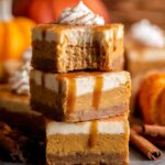 Stacked slices of Pumpkin Cheesecake Bars with a caramel drizzle and a dollop of whipped cream on top, showcasing their creamy texture.