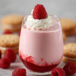 A glass filled with pink Raspberry Mousse, topped with a fresh raspberry and whipped cream. The mousse has a layer of raspberry coulis at the bottom, and is surrounded by fresh raspberries and cookies.