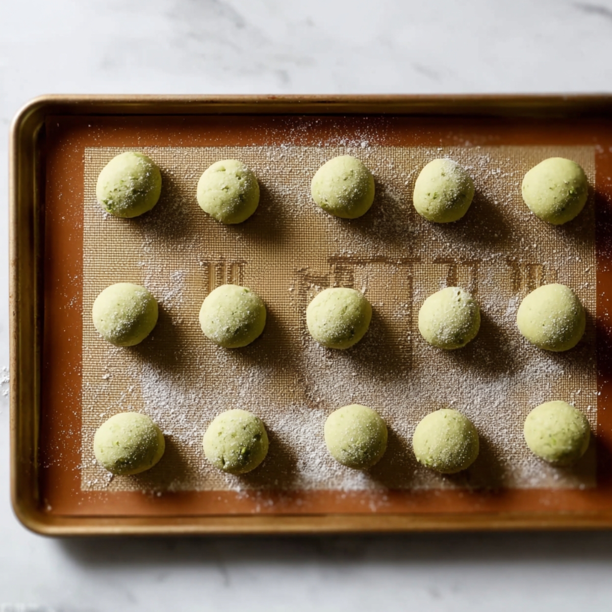 Best Pistachio Drop Cookies Recipe 10 A top-down view of pistachio cookie dough balls, evenly spaced on a silicone-lined baking sheet, ready for baking. The dough balls are lightly dusted with flour.