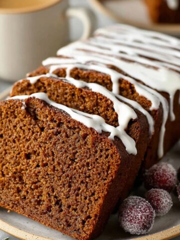 Easy Spiced Gingerbread Loaf Recipe 1 A Spiced Gingerbread Loaf, sliced and topped with a glossy white icing drizzle, with frosted cranberries on the side and a cup of coffee in the background.