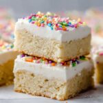Close-up of Sugar Cookie Bars topped with smooth white frosting and colorful sprinkles, stacked neatly on parchment paper.
