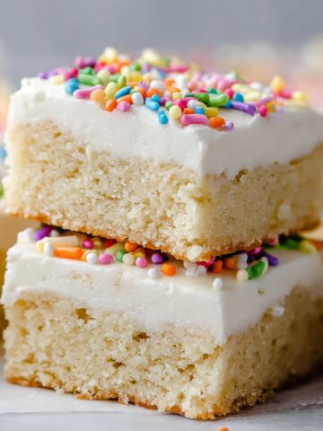 Close-up of Sugar Cookie Bars topped with smooth white frosting and colorful sprinkles, stacked neatly on parchment paper.