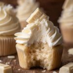 Close-up of a White Chocolate Cupcakes with buttercream frosting topped with a piece of white chocolate. A bite has been taken out, showing the soft, fluffy interior of the cupcake.