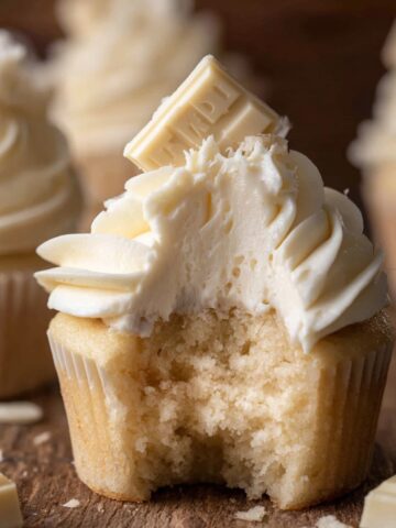 Homemade White Chocolate Cupcakes 2 Close-up of a White Chocolate Cupcakes with buttercream frosting topped with a piece of white chocolate. A bite has been taken out, showing the soft, fluffy interior of the cupcake.