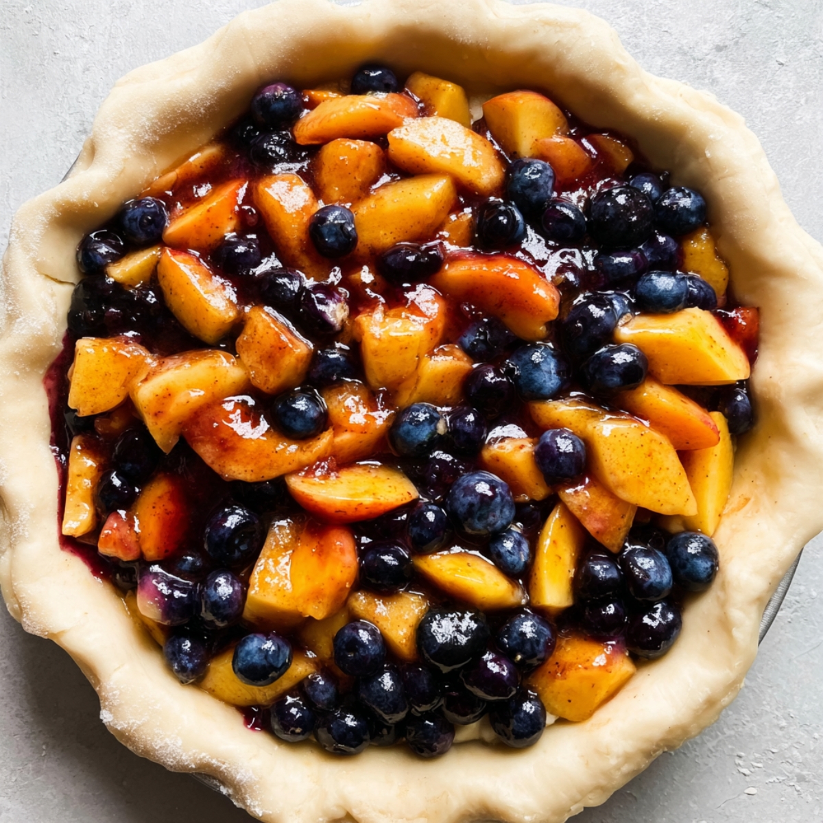 Easy Blueberry Peach Pie Recipe 10 Close-up of a blueberry and peach pie being filled with juicy fruit mixture in an unbaked pie crust, showing vibrant blueberries and peach chunks coated in cinnamon-spiced sauce.