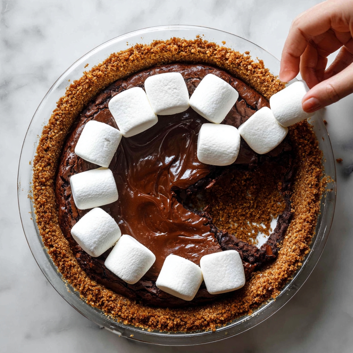 Easy S'mores Brownie Pie Recipe 11 Overhead view of chocolate brownie pie in a graham cracker crust with marshmallows being added by hand before baking.
