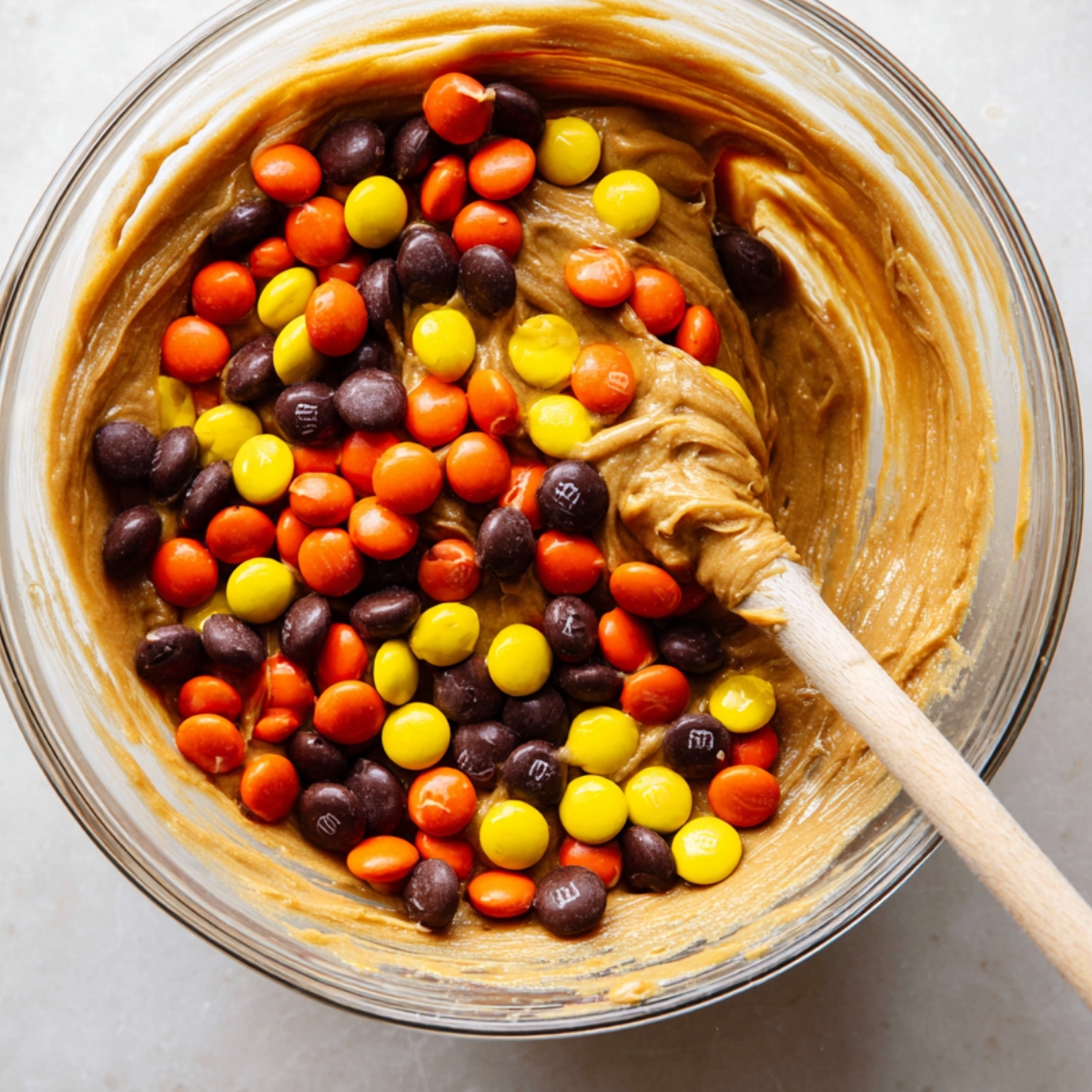 Easy Peanut Butter Blondies Recipe 11 A glass bowl of smooth peanut butter blondie batter, filled with orange, yellow, and brown candy-coated chocolate pieces, with a wooden spatula.