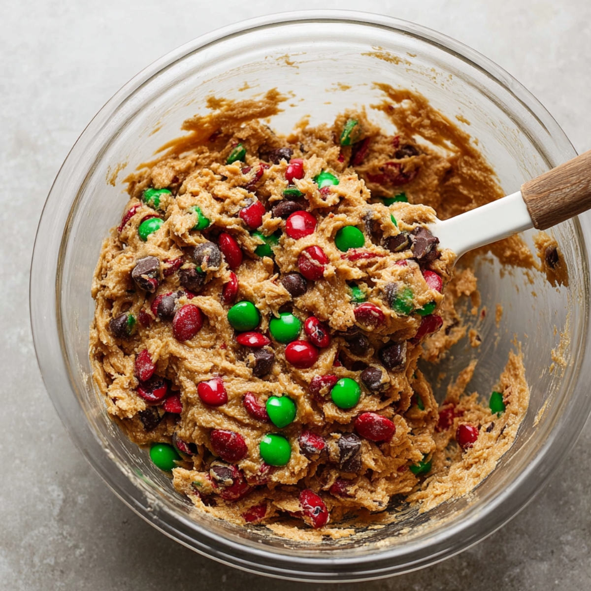 Easy Flourless Monster Cookies Recipe 10 Mixing bowl filled with flourless monster cookie dough with red and green M&M’s, chocolate chips, and oats, being stirred with a spatula.