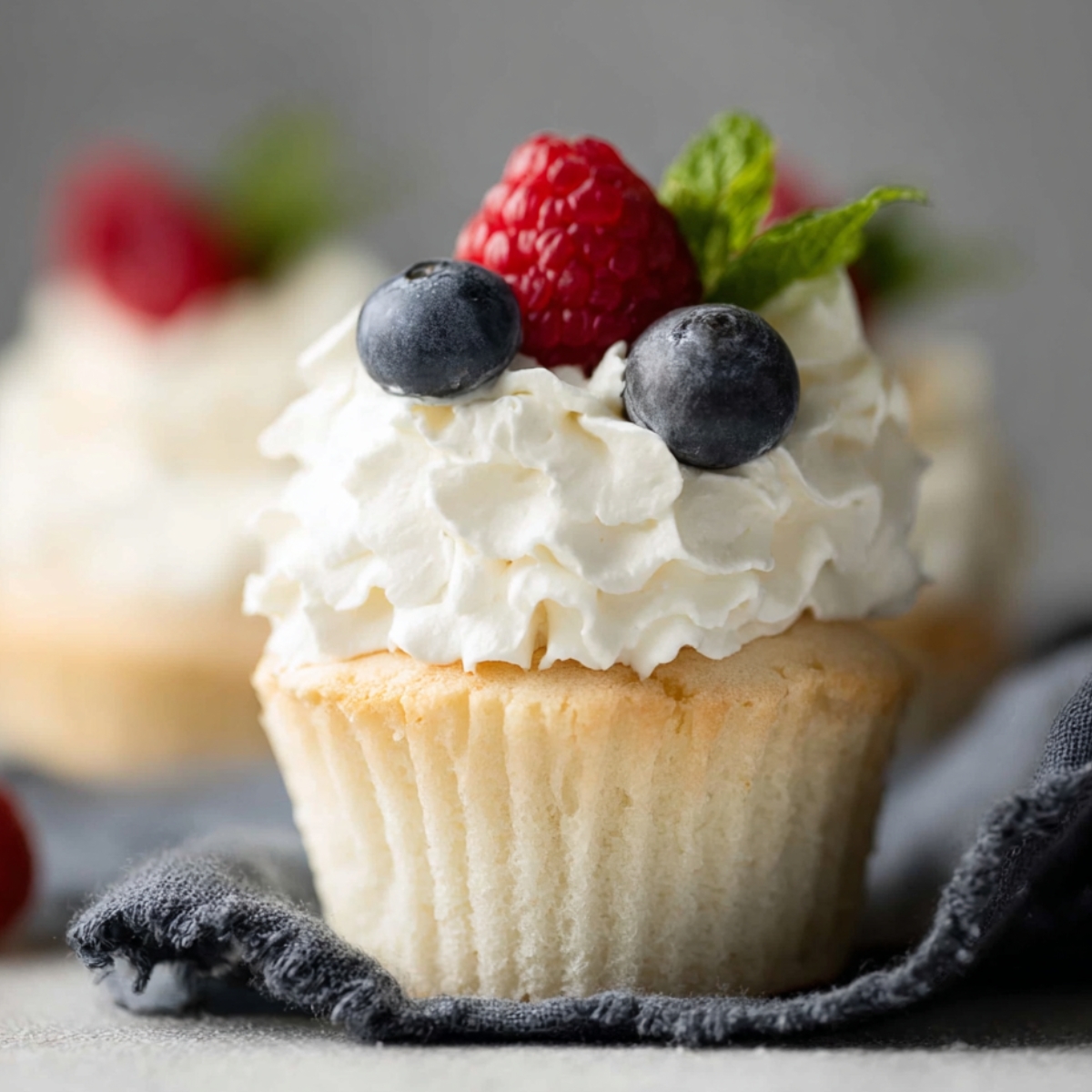Angel Food Cupcakes topped with fluffy whipped cream, fresh raspberries, blueberries, and mint leaves in elegant bakery-style photography.