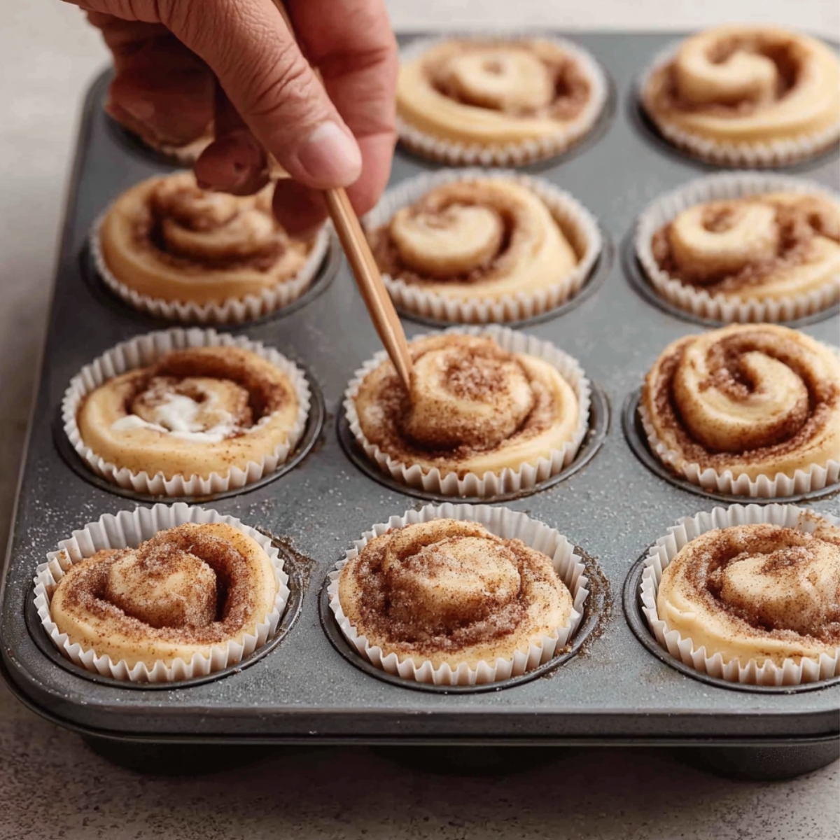 Snickerdoodle Cupcakes with Cinnamon Swirl Frosting 11 A hand using a wooden skewer to swirl cinnamon sugar into the batter in a muffin tin with paper liners. The cinnamon rolls are uncooked and arranged in neat rows.