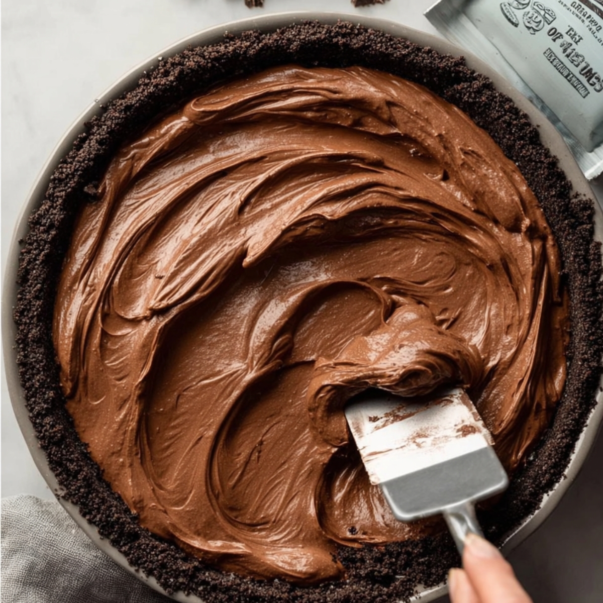 The Best Nutella Cream Pie Recipe 11 A spatula spreading smooth chocolate mousse filling into an Oreo crust pie dish, with the creamy mousse texture clearly visible, and a portion of the crust around the edges.