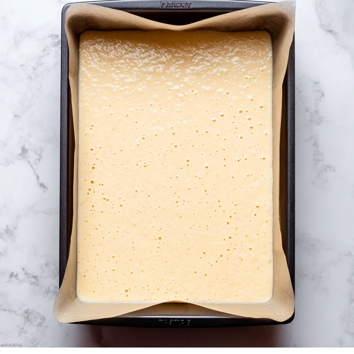 Easy Champagne Cake Roll Recipe 11 Vanilla sponge cake batter spread evenly in a parchment-lined baking pan on a white marble surface before baking.