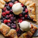 A rustic mixed Berry Galette with a golden, flaky crust, filled with blueberries, raspberries, and blackberries, topped with scoops of vanilla ice cream and a metal spatula lifting a slice.