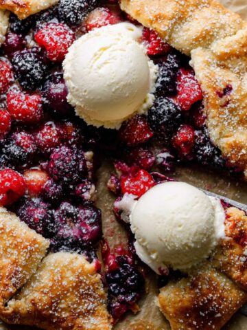 The Best Berry Galette Recipe 1 A rustic mixed Berry Galette with a golden, flaky crust, filled with blueberries, raspberries, and blackberries, topped with scoops of vanilla ice cream and a metal spatula lifting a slice.