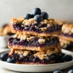 Stack of homemade Blueberry Pie Bars with buttery crumble topping and fresh blueberries on a white plate, rustic bakery-style dessert photography.