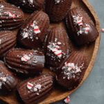 Chocolate Peppermint Madeleines dipped in dark chocolate and sprinkled with crushed peppermint candy, arranged on a wooden plate, festive holiday dessert