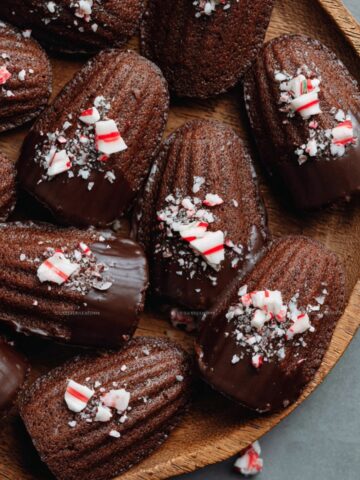 Best Chocolate Peppermint Madeleines 1 Chocolate Peppermint Madeleines dipped in dark chocolate and sprinkled with crushed peppermint candy, arranged on a wooden plate, festive holiday dessert
