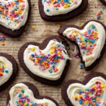 A close-up of decorated heart-shaped Chocolate Sugar Cookies, with thick white frosting topped with colorful sprinkles. One cookie is partially bitten, revealing the soft texture inside.