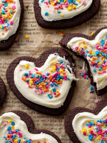Cut-Out Chocolate Sugar Cookies 1 A close-up of decorated heart-shaped Chocolate Sugar Cookies, with thick white frosting topped with colorful sprinkles. One cookie is partially bitten, revealing the soft texture inside.