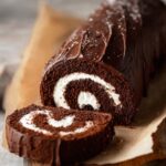 Sliced homemade Chocolate Swiss Roll Cake with a swirl of white cream filling, covered in smooth chocolate ganache, placed on parchment paper on a wooden surface.