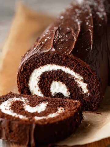 Easy Chocolate Swiss Roll Cake Recipe 1 Sliced homemade Chocolate Swiss Roll Cake with a swirl of white cream filling, covered in smooth chocolate ganache, placed on parchment paper on a wooden surface.