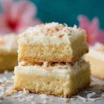 Two stacked Coconut Cream Bars dessert bars with creamy frosting and toasted coconut flakes on top, set on a light surface with soft pastel background for a bright, inviting look.