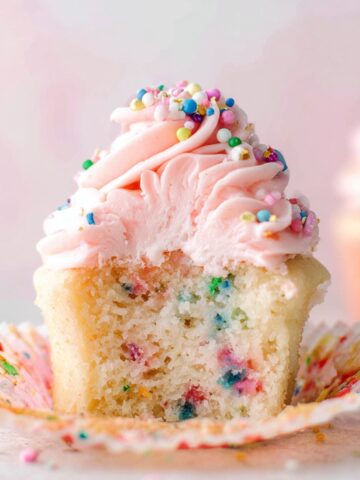 Easy Confetti Sprinkle Cupcakes Recipe 2 Confetti Sprinkle Cupcakes topped with pink buttercream frosting and colorful sprinkles, shown in a partially unwrapped cupcake liner.